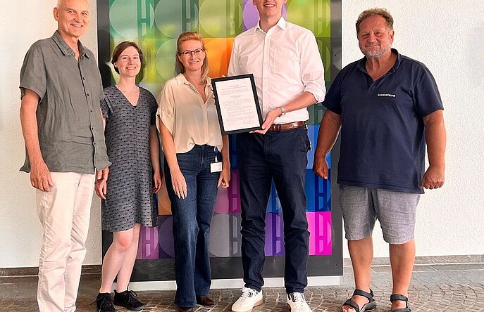Hohenstein EMAS Registration Team Five people stand in front of a wall patterned with colored letter H tiles. The man in the center holds a framed certificate.