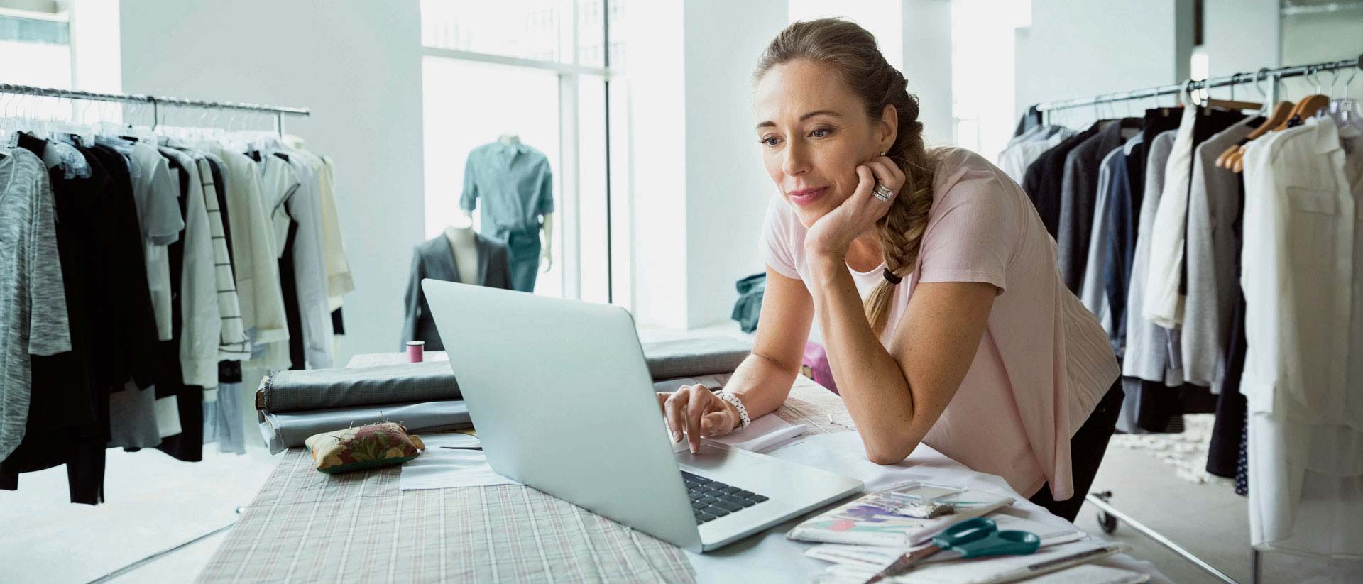 apparel designer on computer in room with with racks of clothes